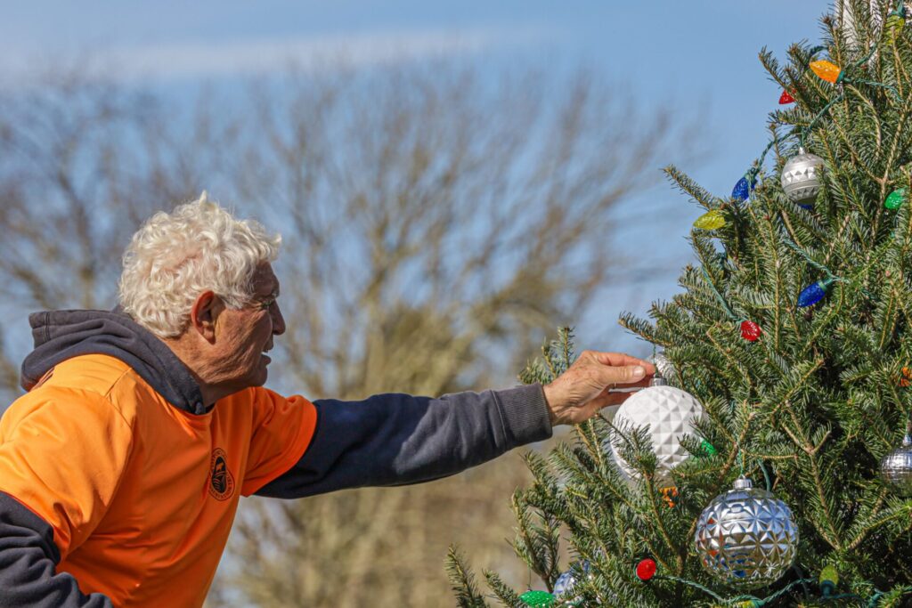 man with white hair and orange t-shirt over a blue hoodie places a sparkly white ornament on a green tree with blue sky