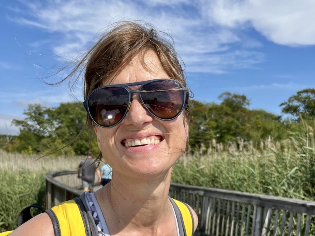 a woman with big sunglasses wearing a backpack smiles at the camera on a boardwalk with blue sky and clouds