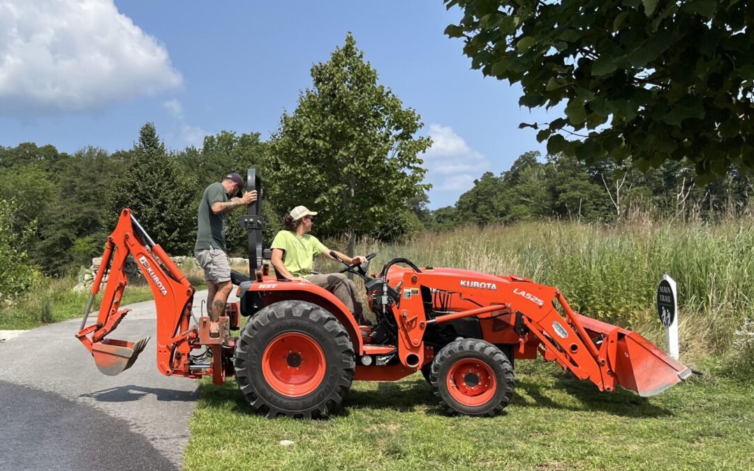 a young man in neon green shirt and ball cap sits on a small orange tractor with a man in a green shirt standing behind